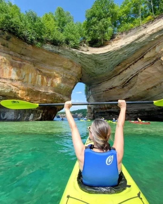 A kayaker raising her paddle under a sandstone arch on turquoise water