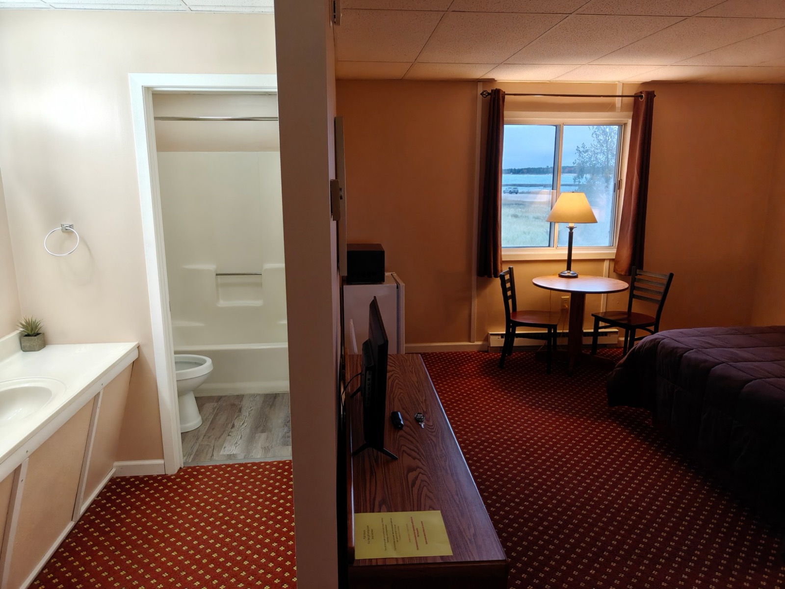 The room from the vanity: a table for two and a lamp by the window looking out on the water, a TV, mini fridge and microwave on the dresser