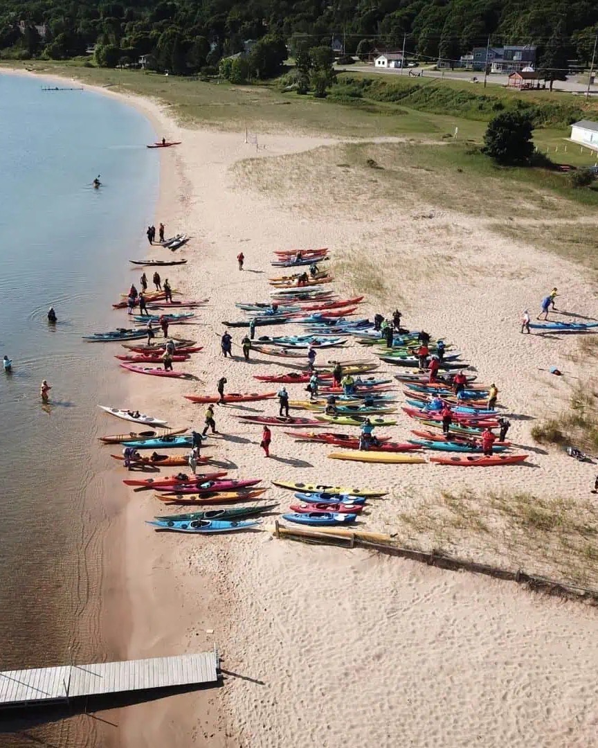 Rows of colorful kayaks on a Grand Marais sand beach from above, paddlers wading into the calm water