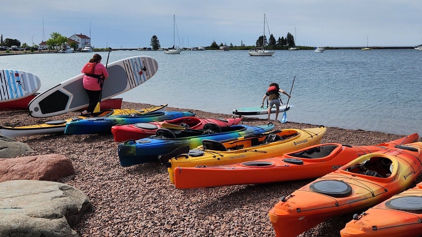 Kayaks and paddleboards on the shore of the Grand Marais harbor, sailboats and the breakwall behind