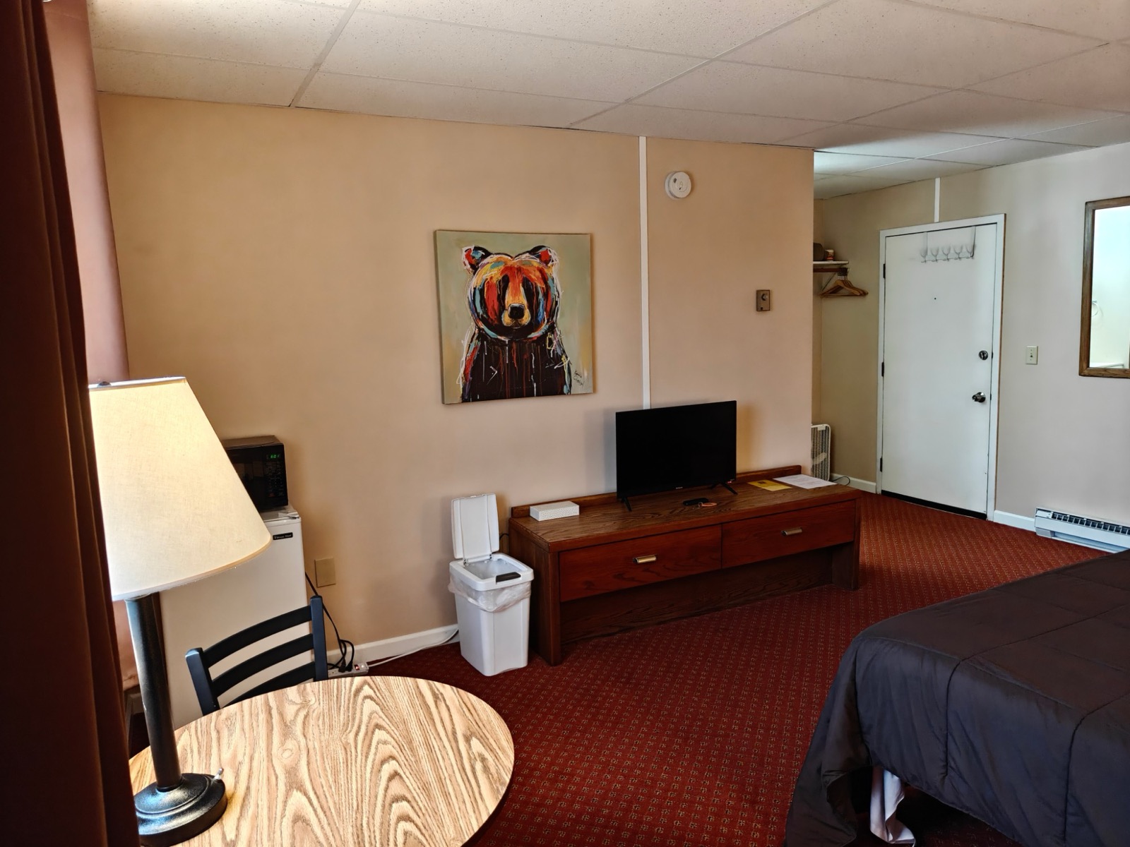 A round table by the window, a TV on the dresser, a mini fridge and microwave under a bear painting